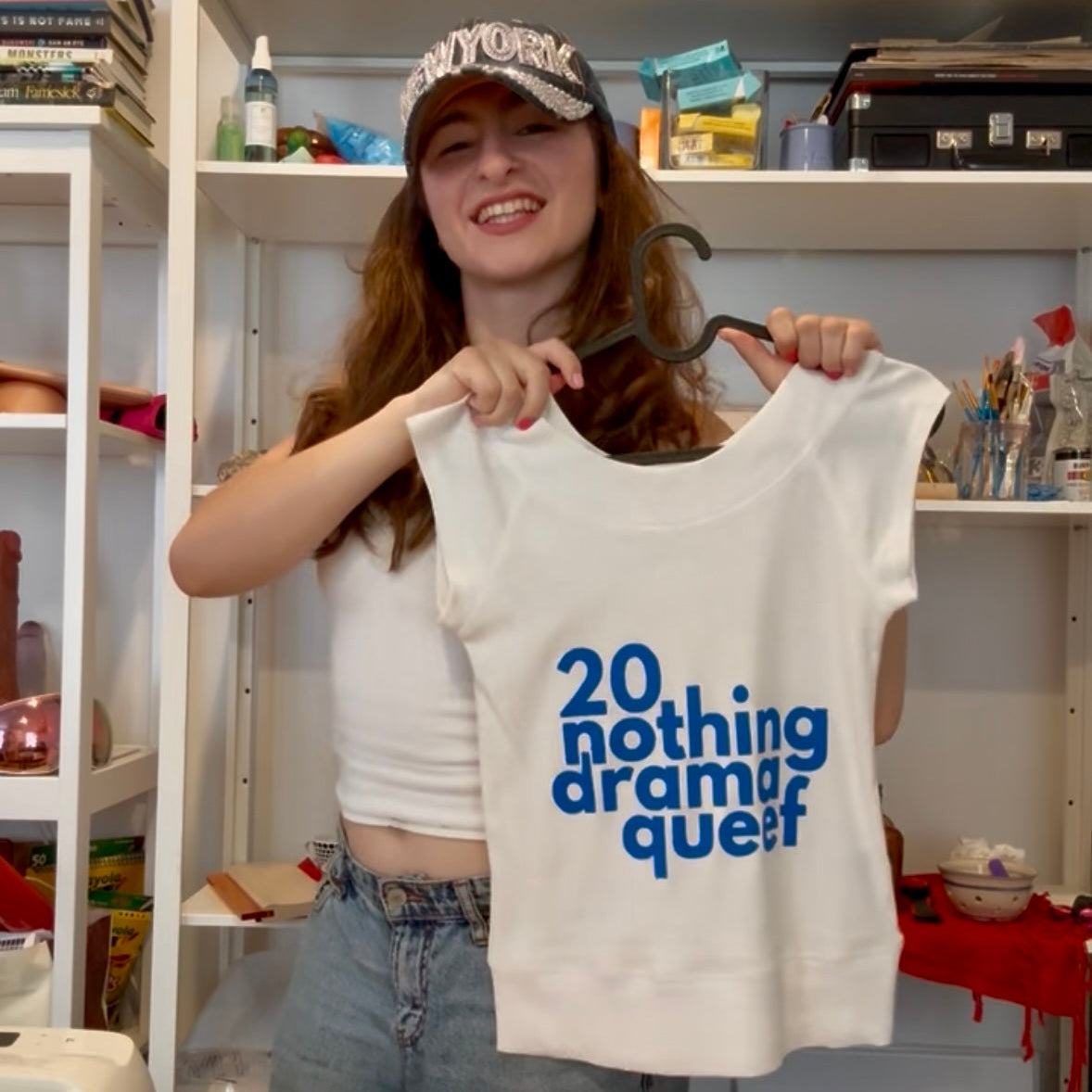 A woman holding up a white sleeveless tank top with blue text that reads '20 nothing drama quest' on a hanger, standing in a room with shelves behind her.