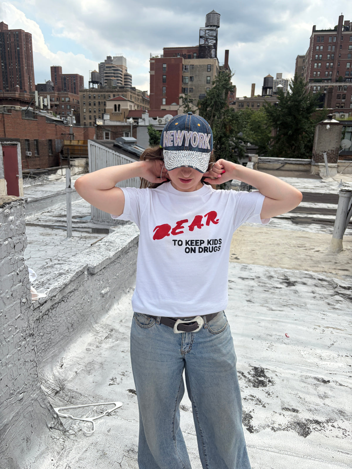 A person standing on a rooftop wearing a white graphic t-shirt with red and black text, blue jeans, and a blue New York cap.