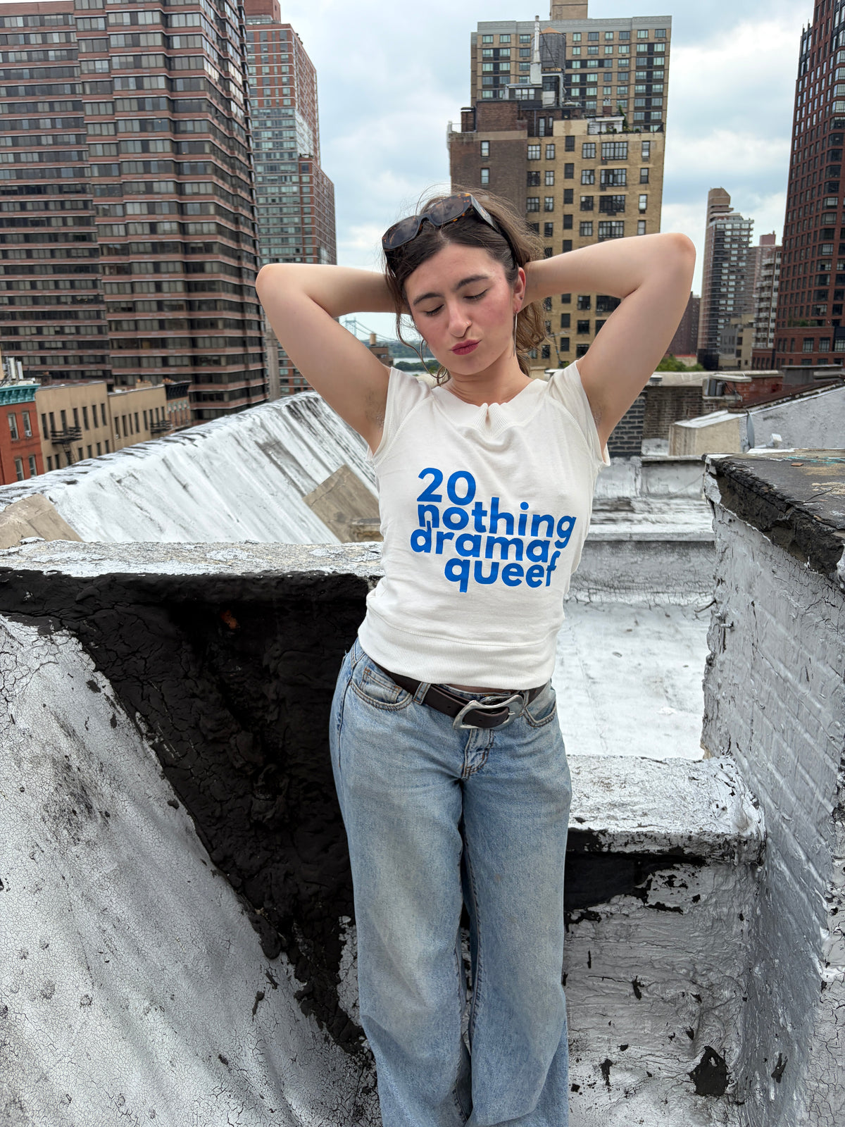 A woman stands on a city rooftop wearing a white graphic T-shirt with blue text reading '20 nothing drama? queer' and light blue jeans, with tall apartment buildings in the background.