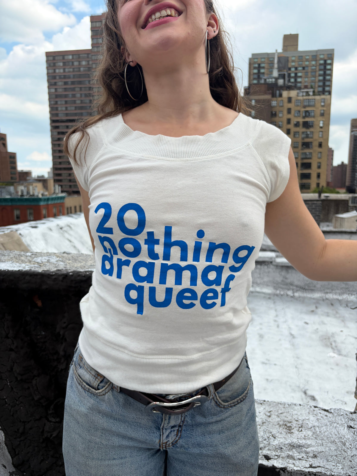 A woman wearing a white scoop-neck t-shirt with blue text that reads '20 nothing drama queer', paired with light-wash blue jeans, photographed outdoors on a rooftop with city buildings in the background.
