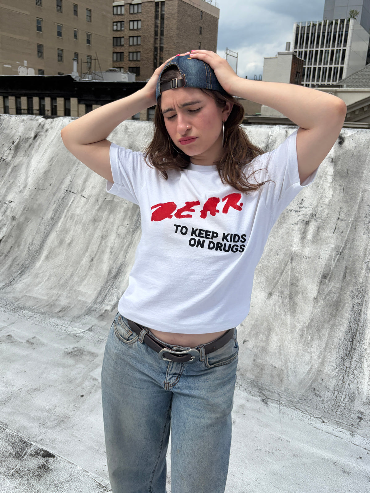 A woman wearing a white graphic t-shirt with red and black text that reads 'DEAR TO KEEP KIDS ON DRUGS', paired with blue jeans and a dark baseball cap, standing outdoors in front of a concrete wall.