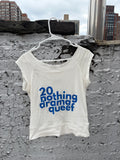An off-white cropped tank top with blue text reading '20 nothing drama queef' hanging on a white plastic hanger against a textured gray concrete wall with city buildings in the background.