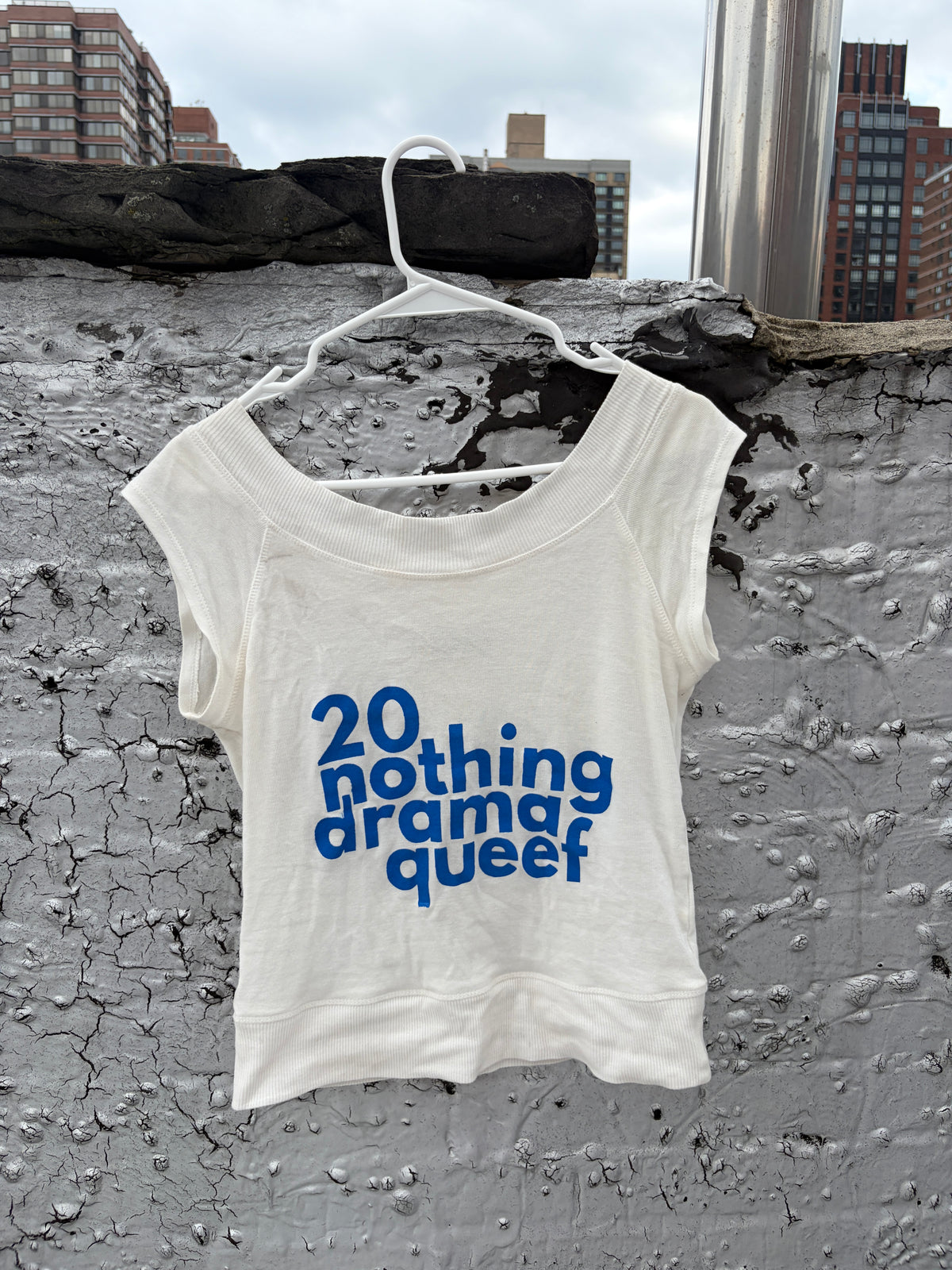 An off-white cropped tank top with blue text reading '20 nothing drama queef' hanging on a white plastic hanger against a textured gray concrete wall with city buildings in the background.