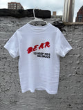 A white short-sleeve t-shirt hanging on a white plastic hanger against a textured gray concrete wall. The shirt features a graphic print with the word 'DEAR' in red brush lettering, and underneath, the phrase 'TO KEEP KIDS ON DRUGS' in black block letters.