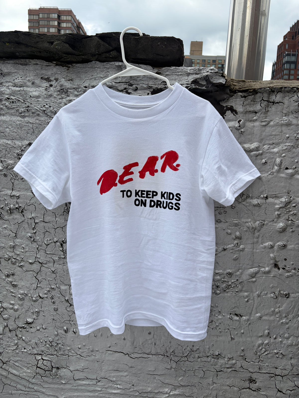 A white short-sleeve t-shirt hanging on a white plastic hanger against a textured gray concrete wall. The shirt features a graphic print with the word 'DEAR' in red brush lettering, and underneath, the phrase 'TO KEEP KIDS ON DRUGS' in black block letters.