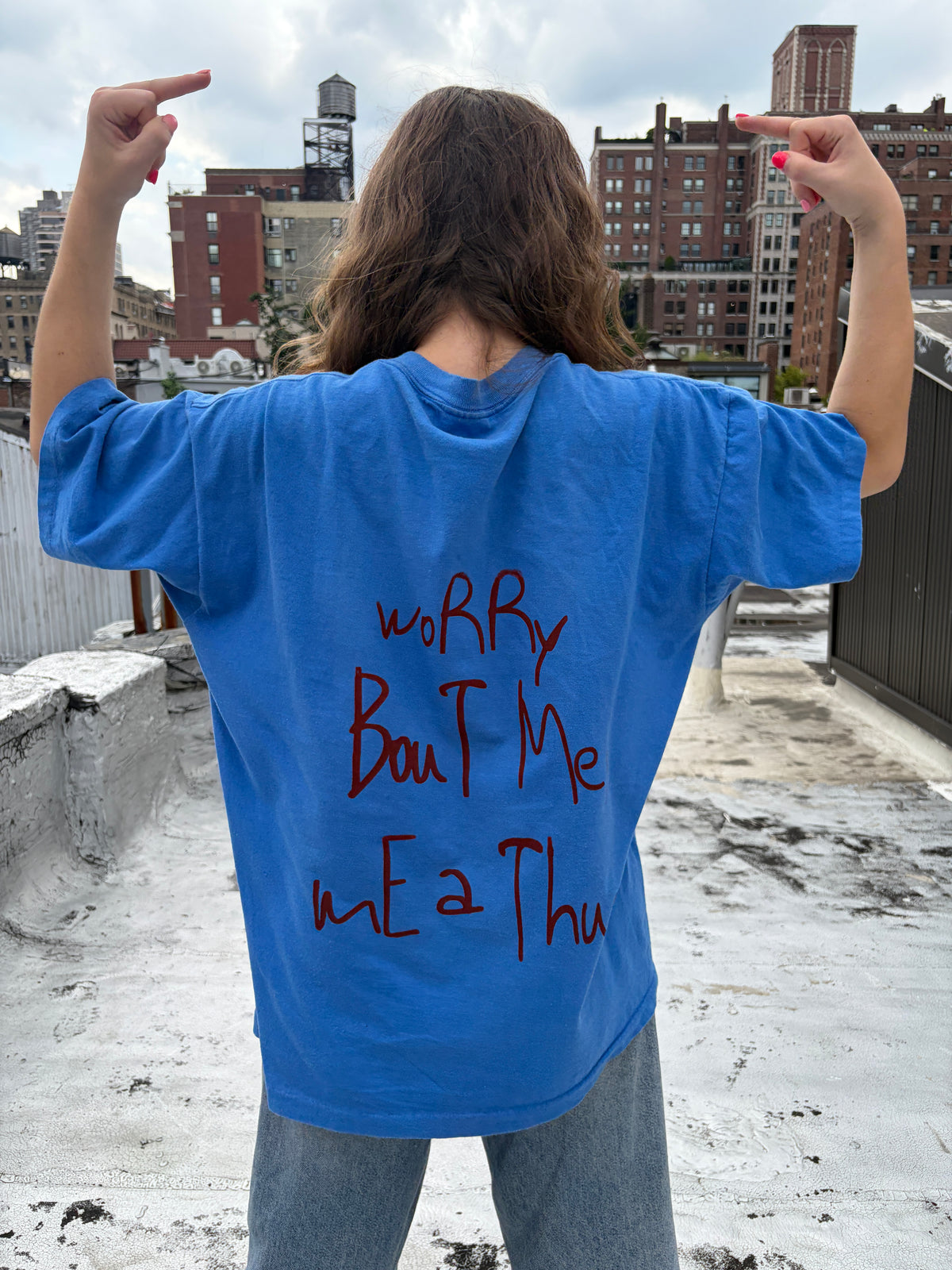 A person viewed from behind wearing a bright blue oversized T-shirt with red handwritten-style text on the back, standing on a rooftop with city buildings in the background.