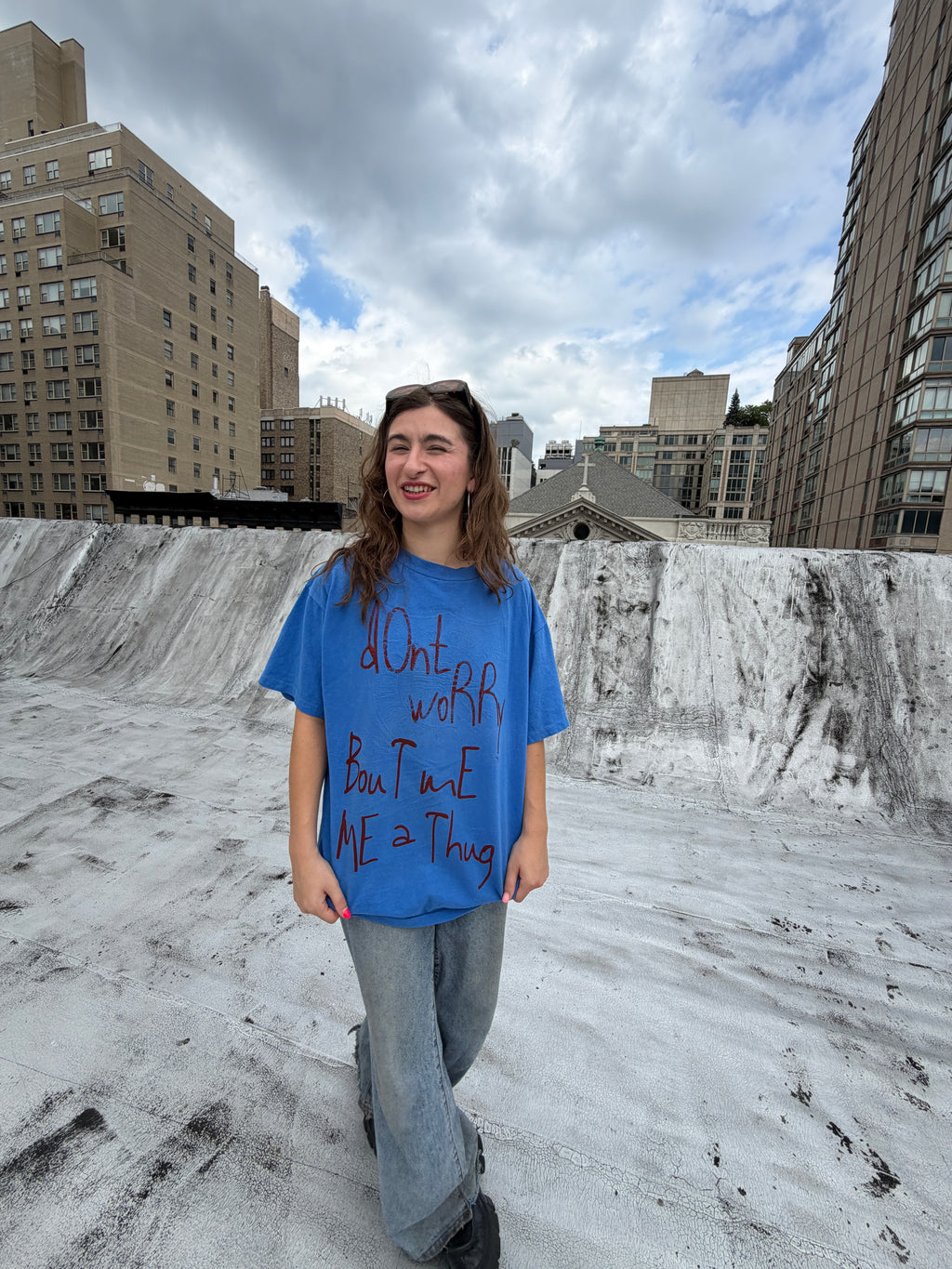 A person stands with arms outstretched on a snowy rooftop, wearing a bright blue graphic T-shirt and light blue jeans, with city buildings and a cloudy sky in the background.