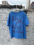 A blue oversized graphic t-shirt hanging on a white plastic hanger against a textured gray concrete wall with city buildings in the background. The shirt features the phrase 'Don't worry But wait ME a Thug' printed in a dark red handwritten-style font.