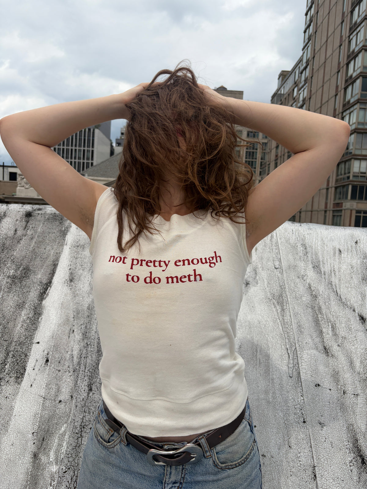A person standing on a rooftop wearing a cream-colored sleeveless t-shirt with red text that reads 'not pretty enough to do meth', paired with blue jeans and a brown belt.