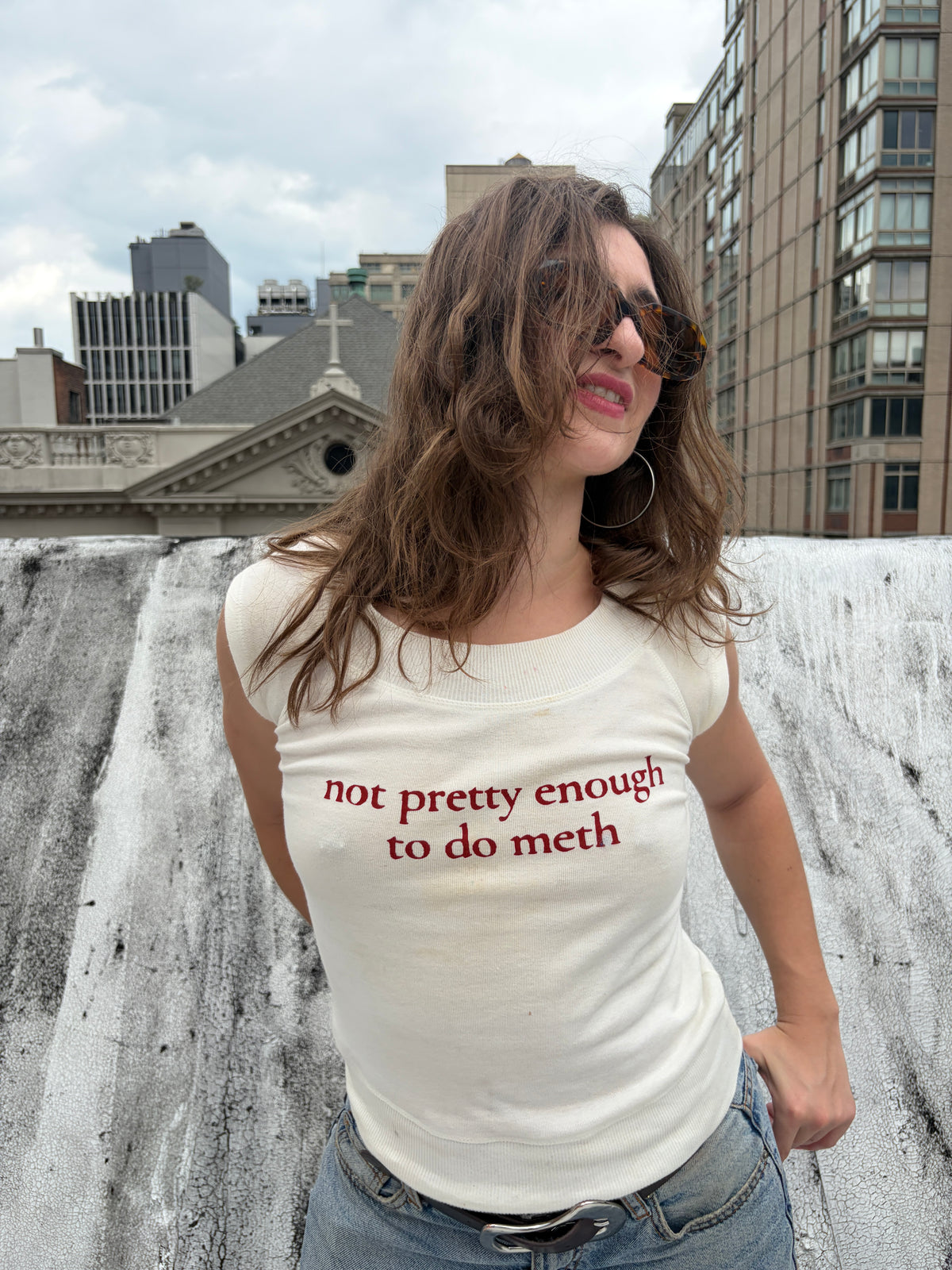 A woman wearing a cream-colored sleeveless t-shirt with the text 'not pretty enough to do meth' printed in red across the front, posing outdoors against a textured concrete wall with city buildings in the background.