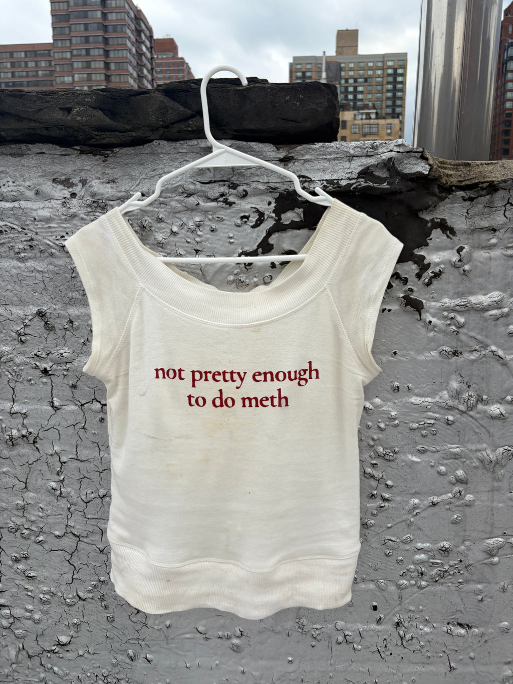 An off-white sleeveless tank top hanging on a white plastic hanger against a textured concrete wall with city buildings in the background. The shirt has red text printed on the front that reads 'not pretty enough to do meth'.