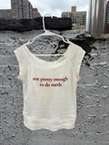 An off-white sleeveless tank top hanging on a white plastic hanger against a textured concrete wall with city buildings in the background. The shirt has red text printed on the front that reads 'not pretty enough to do meth'.