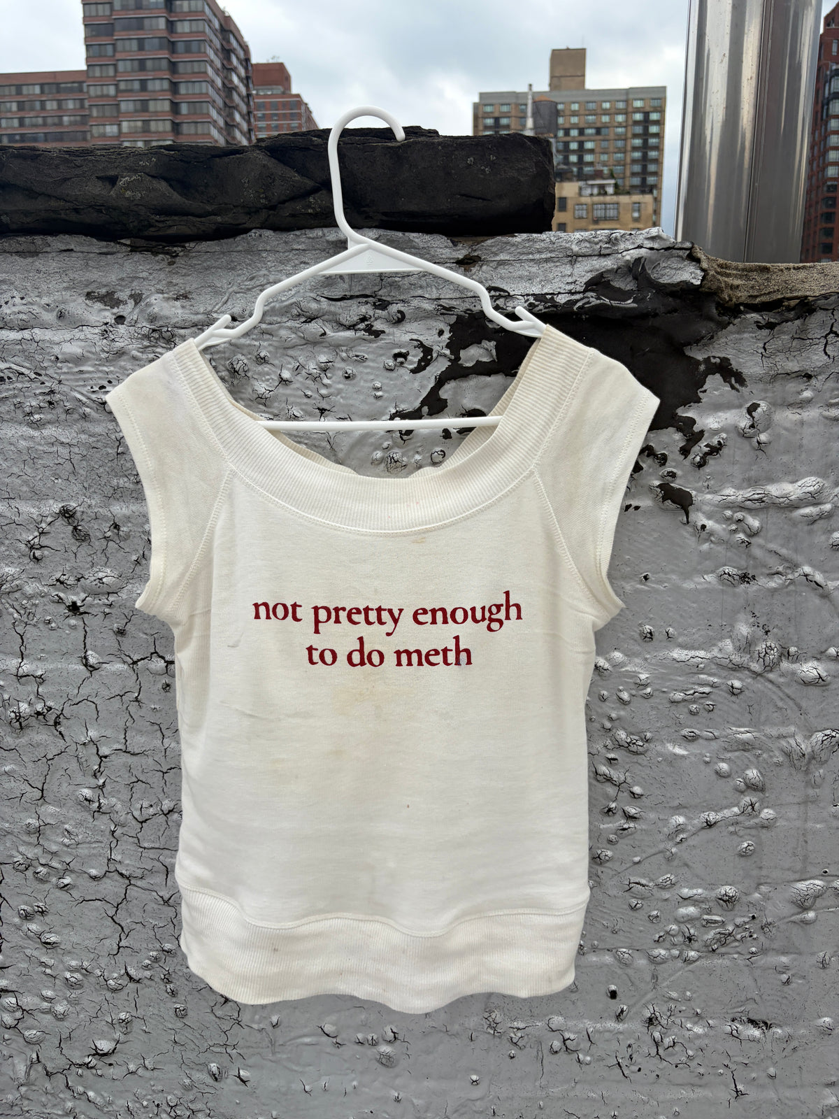 An off-white sleeveless tank top hanging on a white plastic hanger against a textured concrete wall with city buildings in the background. The shirt has red text printed on the front that reads 'not pretty enough to do meth'.