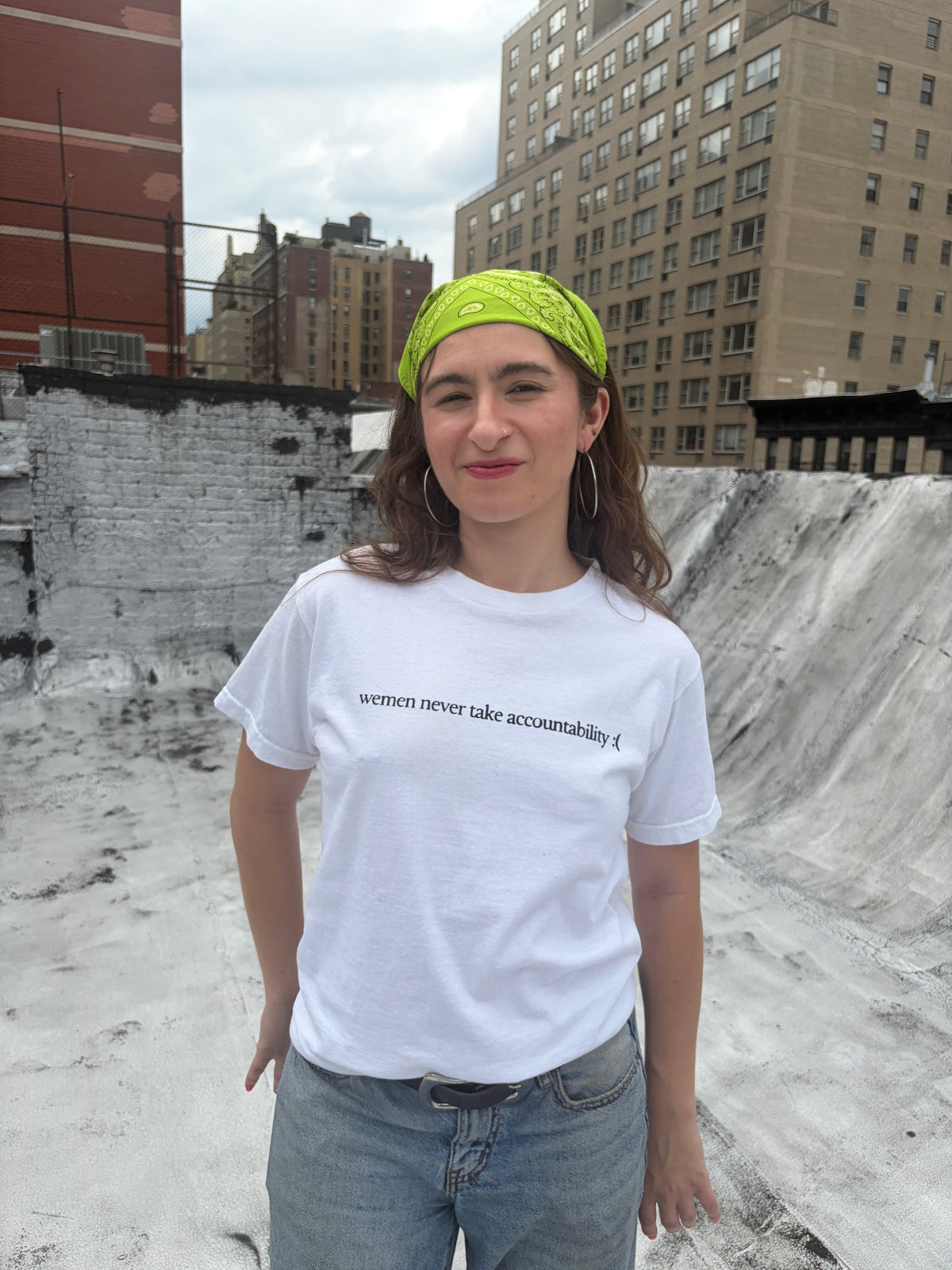 A woman wearing a white graphic t-shirt with the text 'wemen never take accountability :( ' and a bright lime green patterned headband, standing outdoors on a concrete rooftop with city buildings in the background.