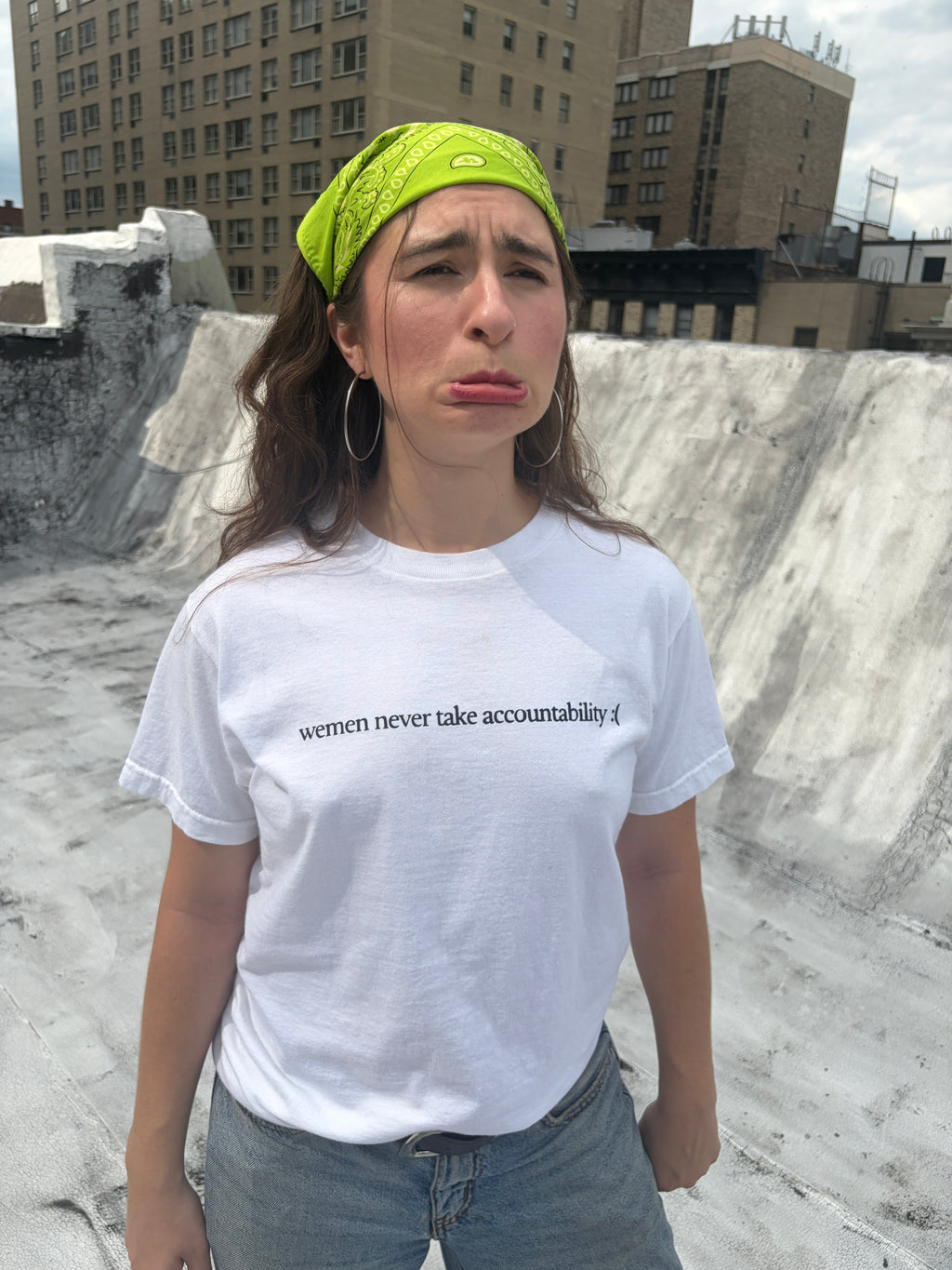 A woman wearing a white t-shirt with the black printed text 'wemen never take accountability :( ' stands on a concrete rooftop with city buildings in the background.