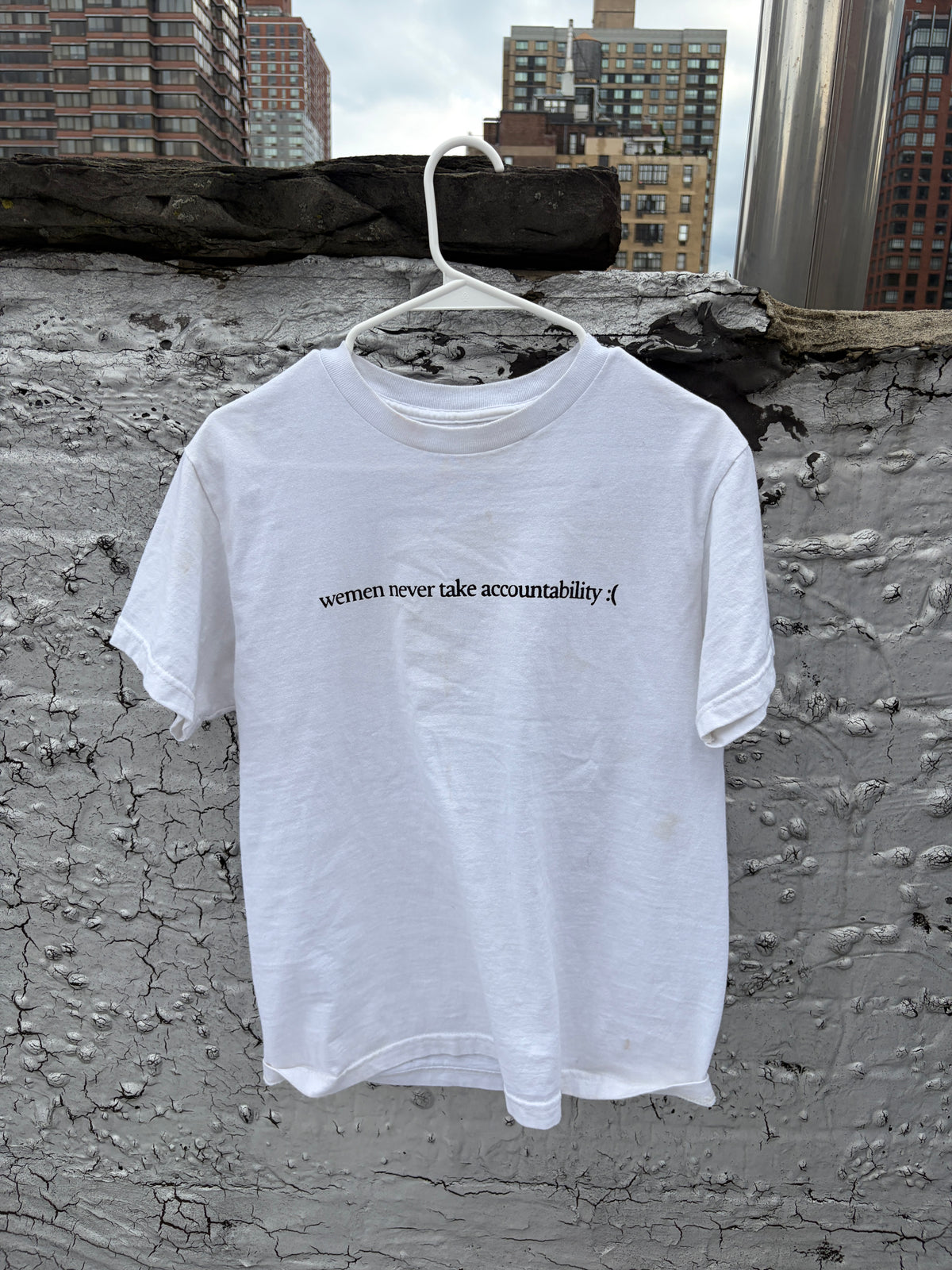 A white short-sleeve t-shirt hanging on a white plastic hanger against a textured concrete wall with city buildings in the background. The shirt features a small black printed text on the chest that reads 'wemen never take accountability :( '